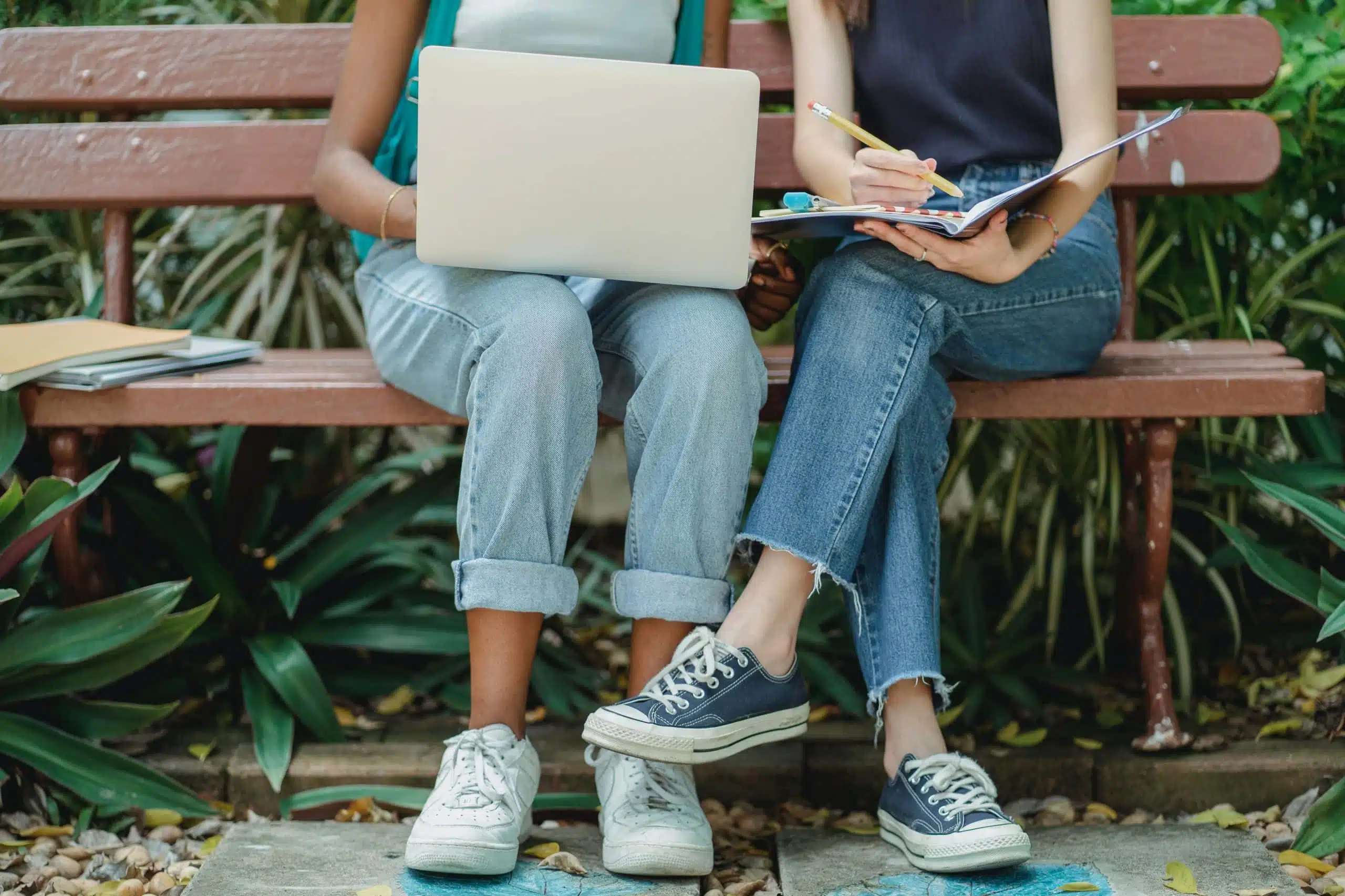 Two students, one holding a laptop, the other a note book, studying together on a bench.